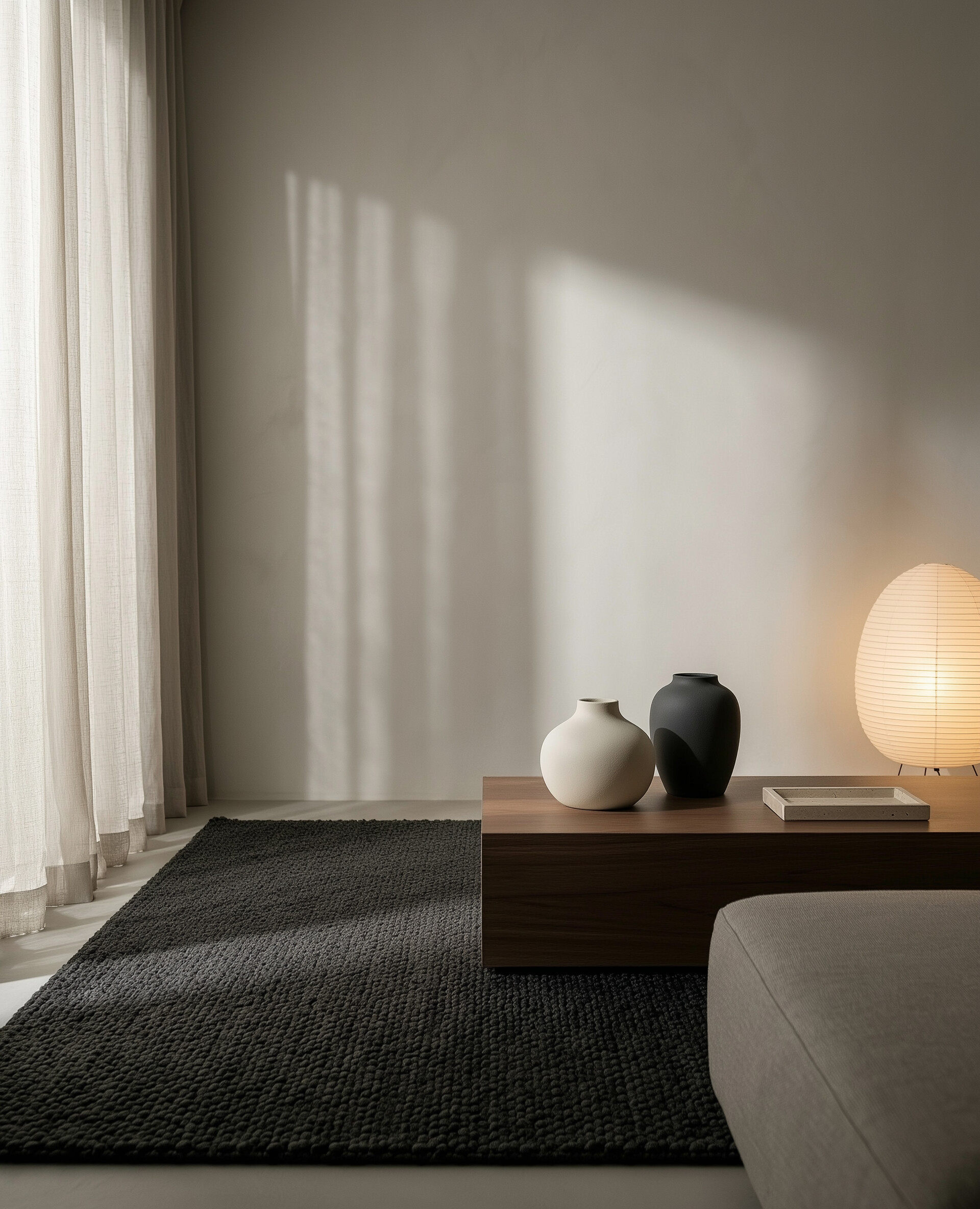 Ambiance japandi minimaliste : tapis anthracite, table basse en bois fumé, vases en céramique mate et lampe en papier, lumière douce et ombres maîtrisées.