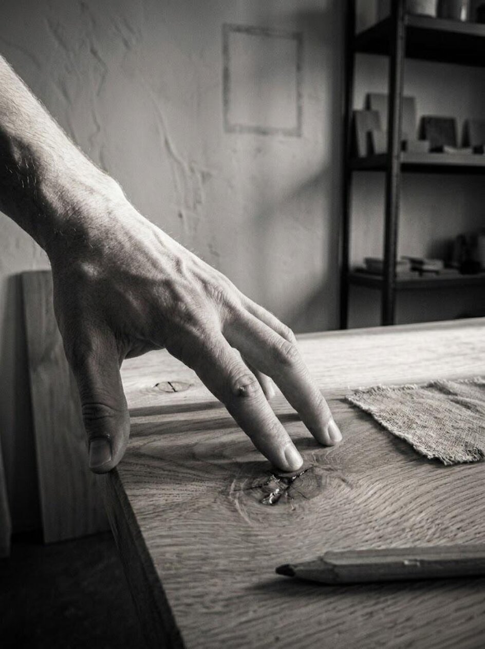 Main effleurant le grain d'un plateau en chêne brut dans un atelier baigné de lumière rasante — geste de sélection, nœud de bois à la résine ambrée, échantillon de lin et crayon de menuisier, noir et blanc
