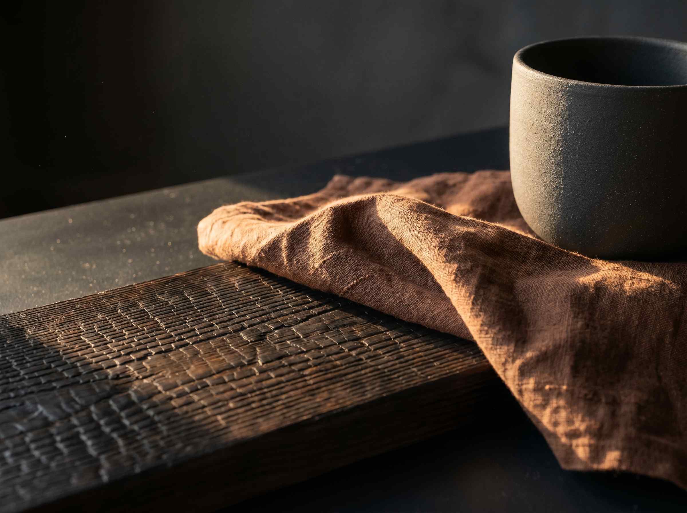 Macro Dark Japandi 2026, surface shou sugi ban en cedre brule, drape de lin chocolat et ceramique anthracite mate sous lumiere rasante