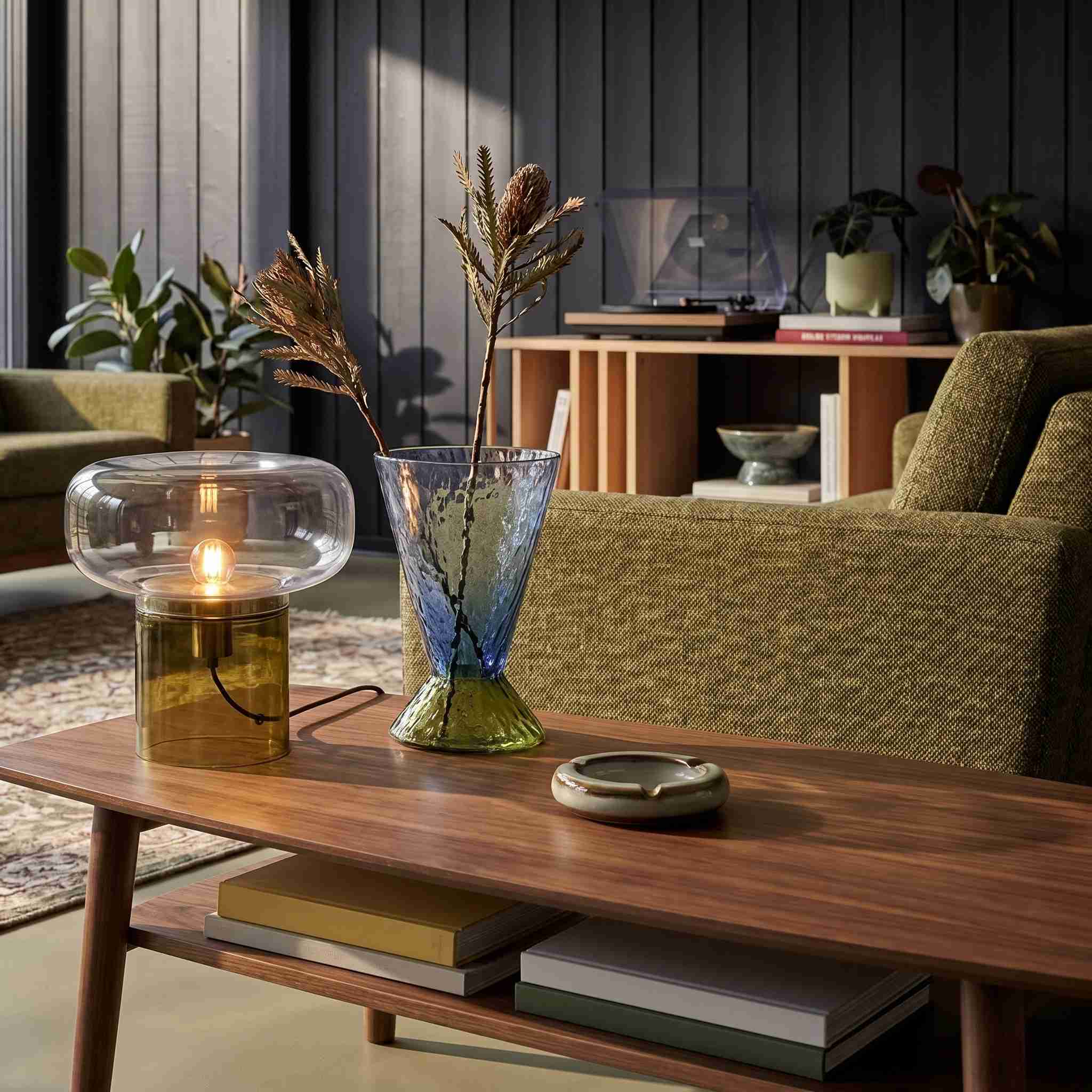 Intérieur Univers Graphique Atelier XVI : table basse noyer, lampe verre ambré, vase verre bleu sur fond panneaux noyer, canapé olive et platine vinyle