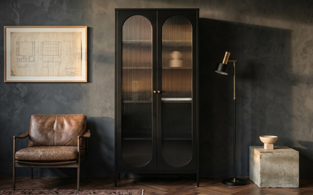 Vitrine colonne en acier laqué noir mat à deux longues arches percées et verre cannelé, salon mature aux murs enduit gris sombre, parquet chevron, fauteuil cuir camel patiné, lampadaire laiton et plinthe béton brut avec bol pierre
