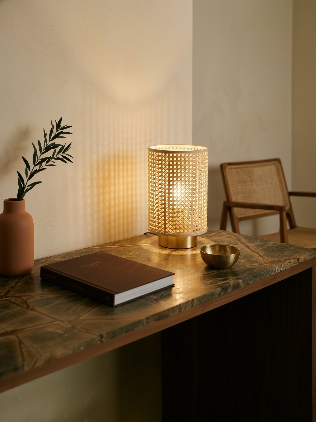 Nature morte contemporaine du rotin retrouvé, lampe à poser à abat-jour cannage allumée diffusant son motif tressé sur le mur de plâtre crème, plateau en marbre veiné brun-vert, vase en terre cuite mate, livre relié et coupelle en laiton bruni, fauteuil cannage flou en arrière-plan