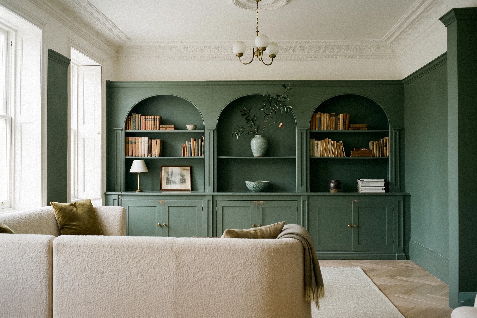 Salon bibliothèque vert sauge dans une maison de ville, boiseries à arches peintes du sol au plafond, canapé en laine bouclée écrue, lustre à globes opalins, corniche moulurée et parquet à chevrons