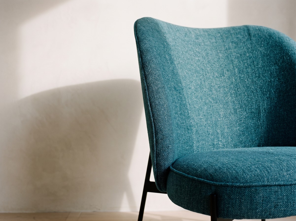 Fauteuil bleu canard au dossier galbé, tissu chiné mat et pieds fuselés noirs, ombre portée sur mur blanc cassé, silhouette sculpturale