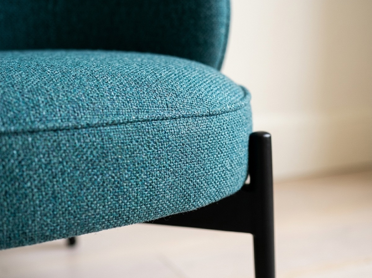 Détail d'un fauteuil bleu canard en tissu chiné mat, assise ronde passepoilée et pied noir fuselé