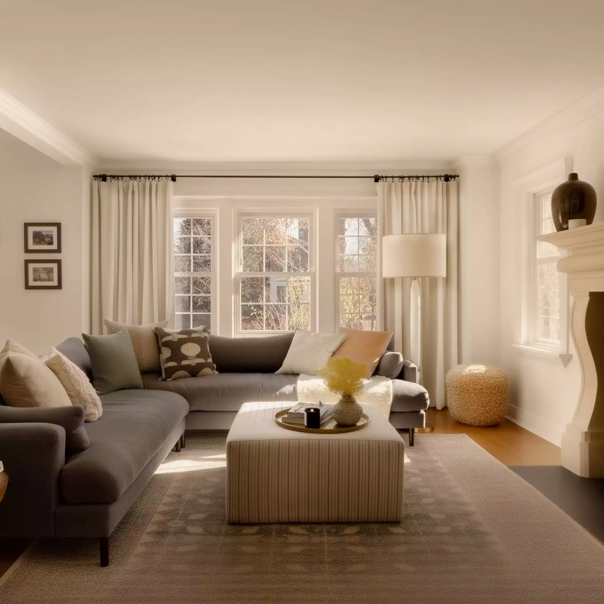 Salon scandinave chaleureux avec grand canapé gris, rideaux beige, ottoman rayé au centre, lampe sur pied ivoire, panier tressé et lumière douce