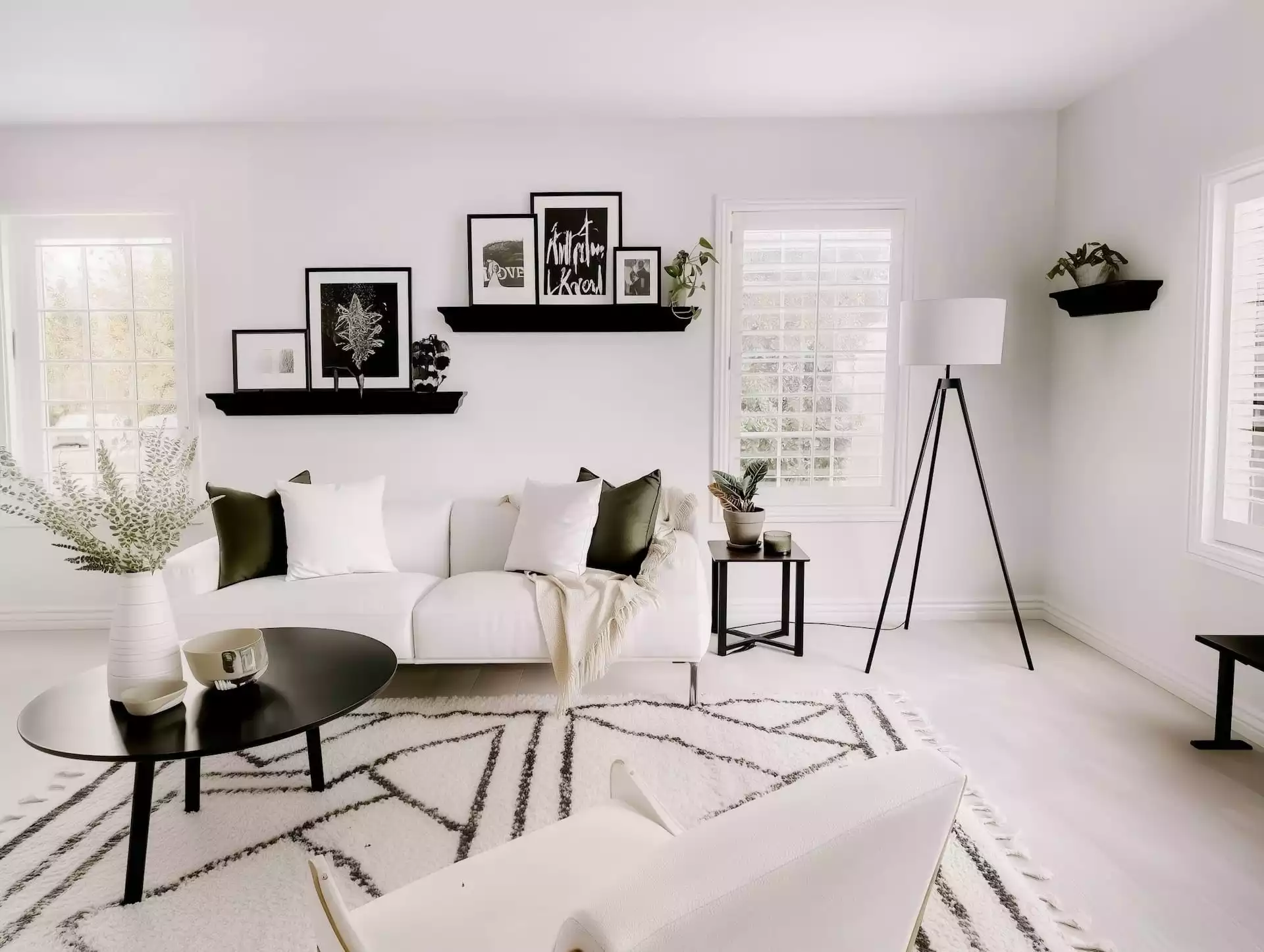 Salon clair avec canapé blanc, étagères galeries noires et cadres, lampadaire trépied noir, tables basses rondes, tapis graphique noir-crème