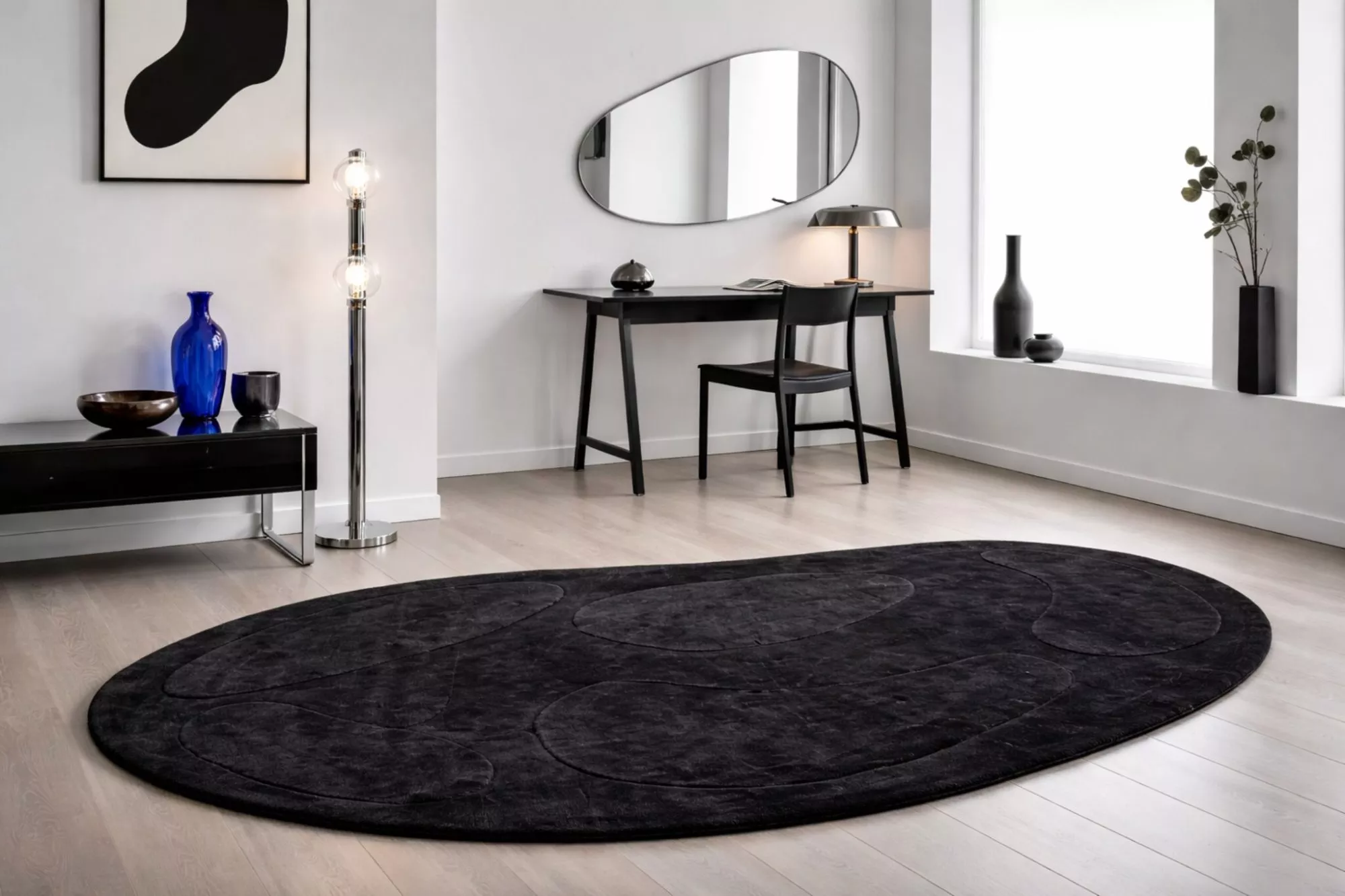 Tapis ovale noir texturé au sol dans un intérieur minimaliste blanc avec bureau noir, miroir organique et lumière naturelle.