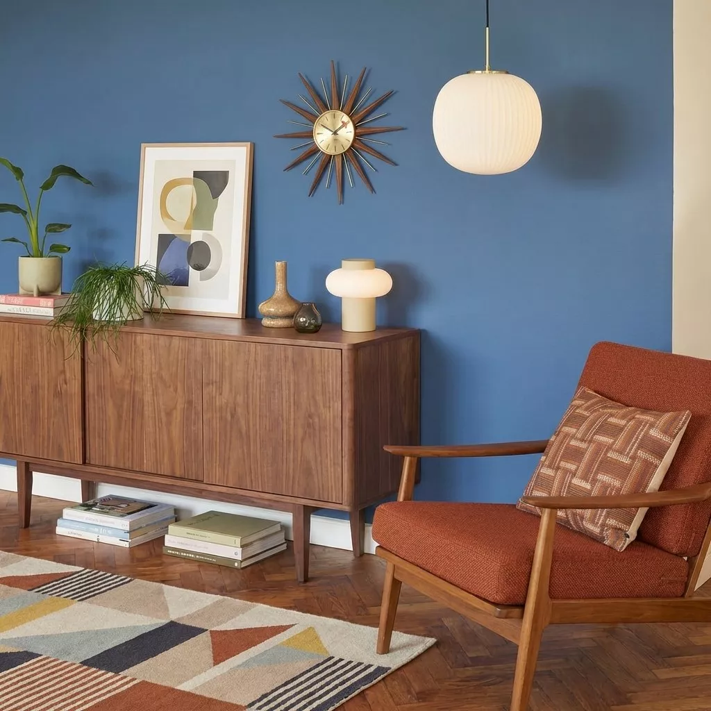 Suspension globe opalin nervuré dans un salon Mid-Century Modern, mur bleu ardoise, buffet noyer, horloge soleil et fauteuil rouille