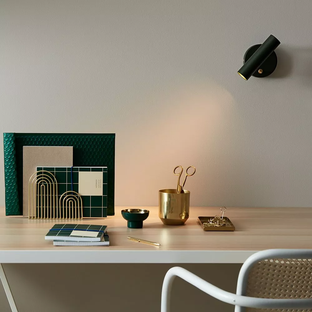 Applique cylindre orientable noir et laiton allumée au-dessus d'un bureau clair, range-revues laiton et carnets verts