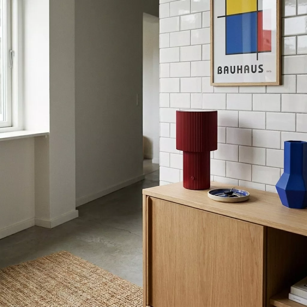 Lampe à poser rechargeable rouge sur un meuble en bois clair, mur en briques blanches et affiche d'art géométrique.