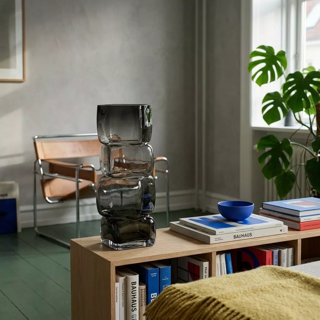 Vase verre fumé en colonne de cubes sur enfilade bois clair, chaise tube et cuir, sol vert et grand feuillage