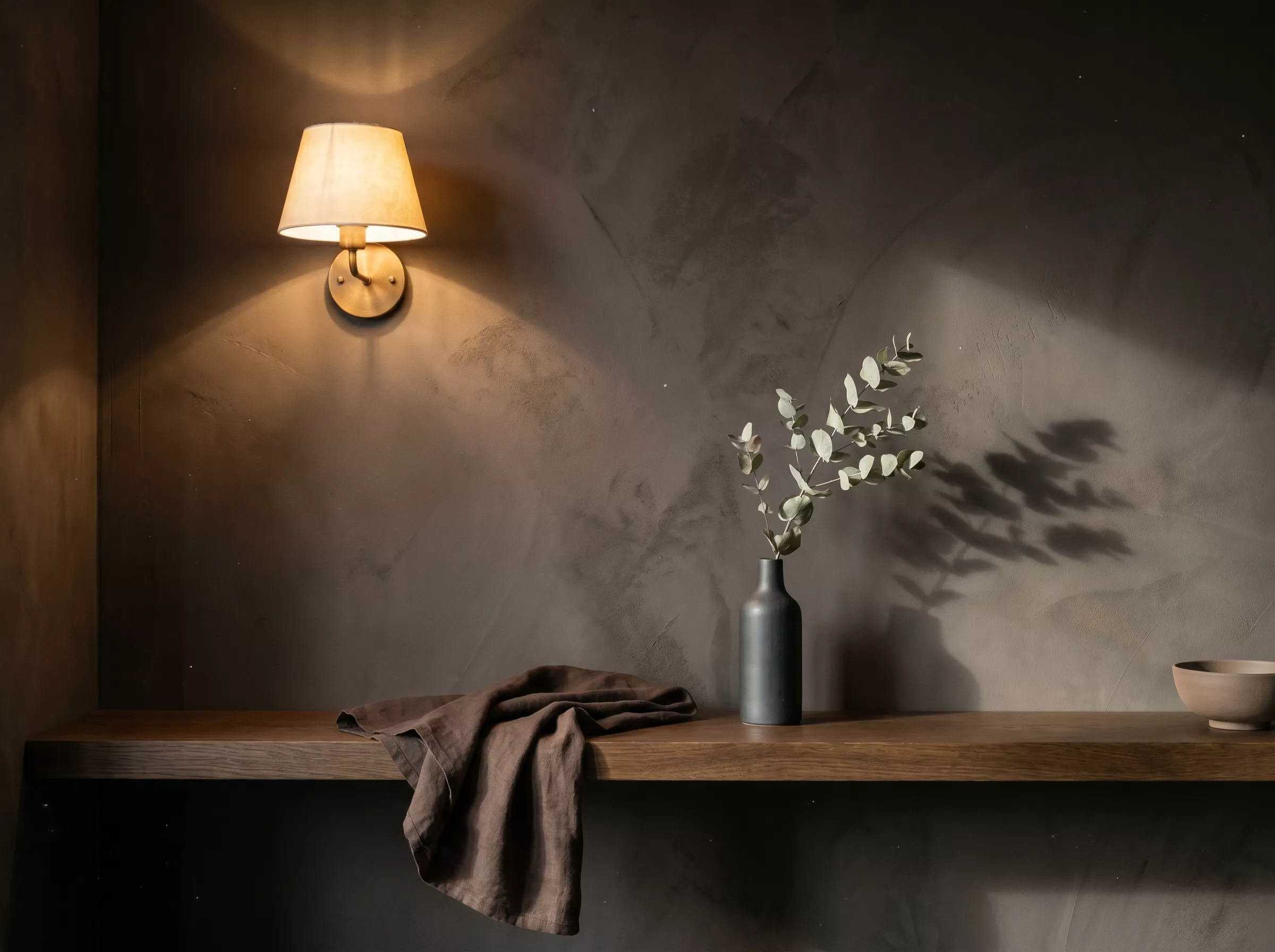 Palette Dark Japandi, mur clay-plaster charcoal mottled, étagère en noyer fumé et vase en grès anthracite avec branche d'eucalyptus sous applique en laiton brossé