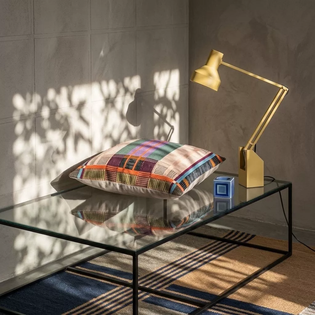 Coussin brodé multicolore motifs géométriques posé sur table basse en métal et verre, mur béton et lampe articulée dorée