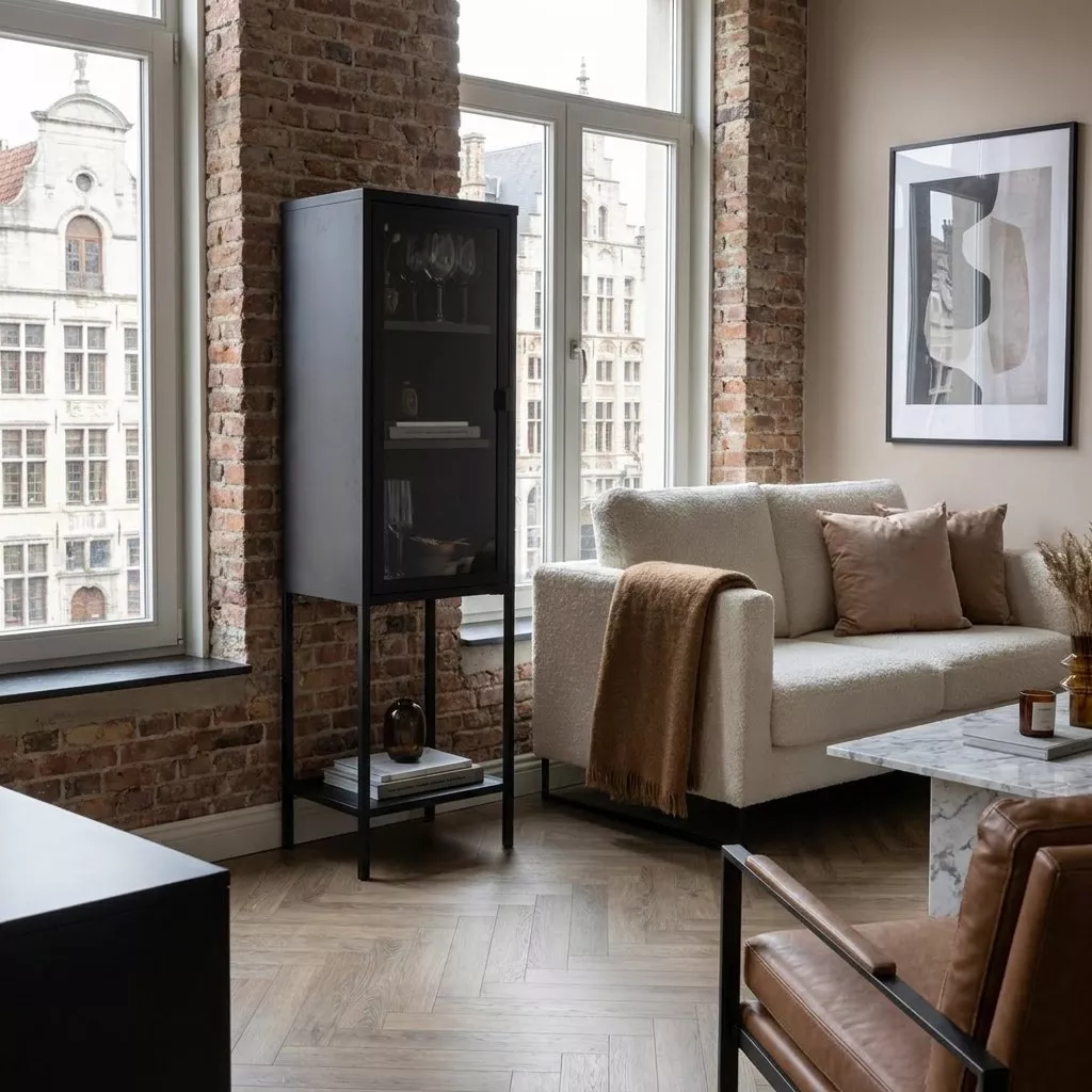 Vitrine colonne noire en acier thermolaqué et verre clair, dressée contre un mur de brique entre deux fenêtres.