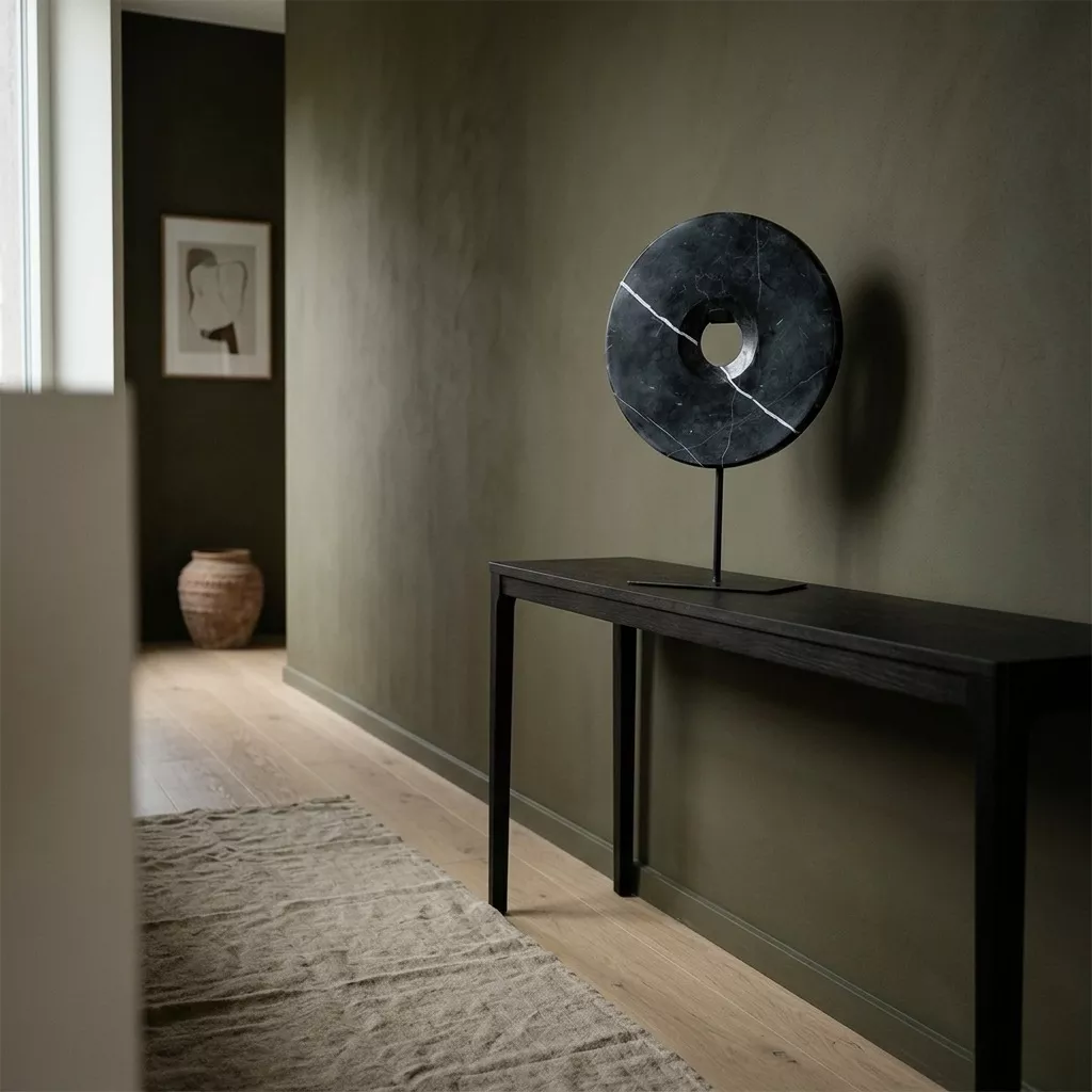 Disque en marbre noir veiné sur console en métal noir, mur enduit olive, sol en parquet clair et tapis en fibre naturelle