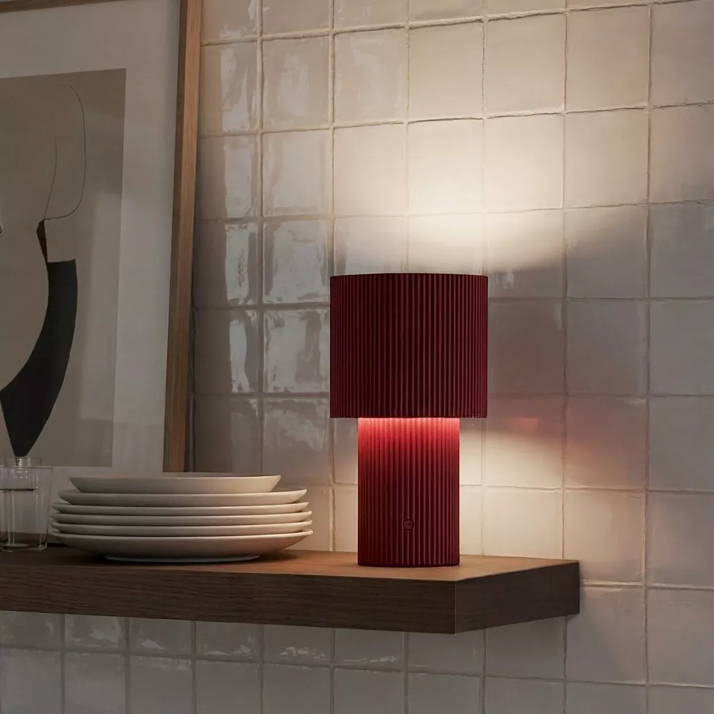 Lampe à poser rechargeable rouge sur une étagère en bois foncé, mur en carrelage artisanal beige et vaisselle empilée.