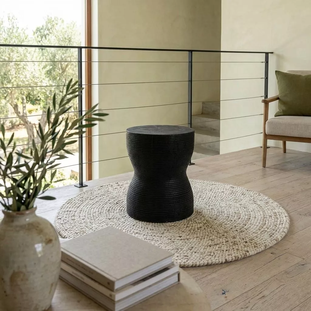 Tabouret en bois massif noir posé sur un tapis rond en jute, près d'une grande plante et de murs clairs lumineux