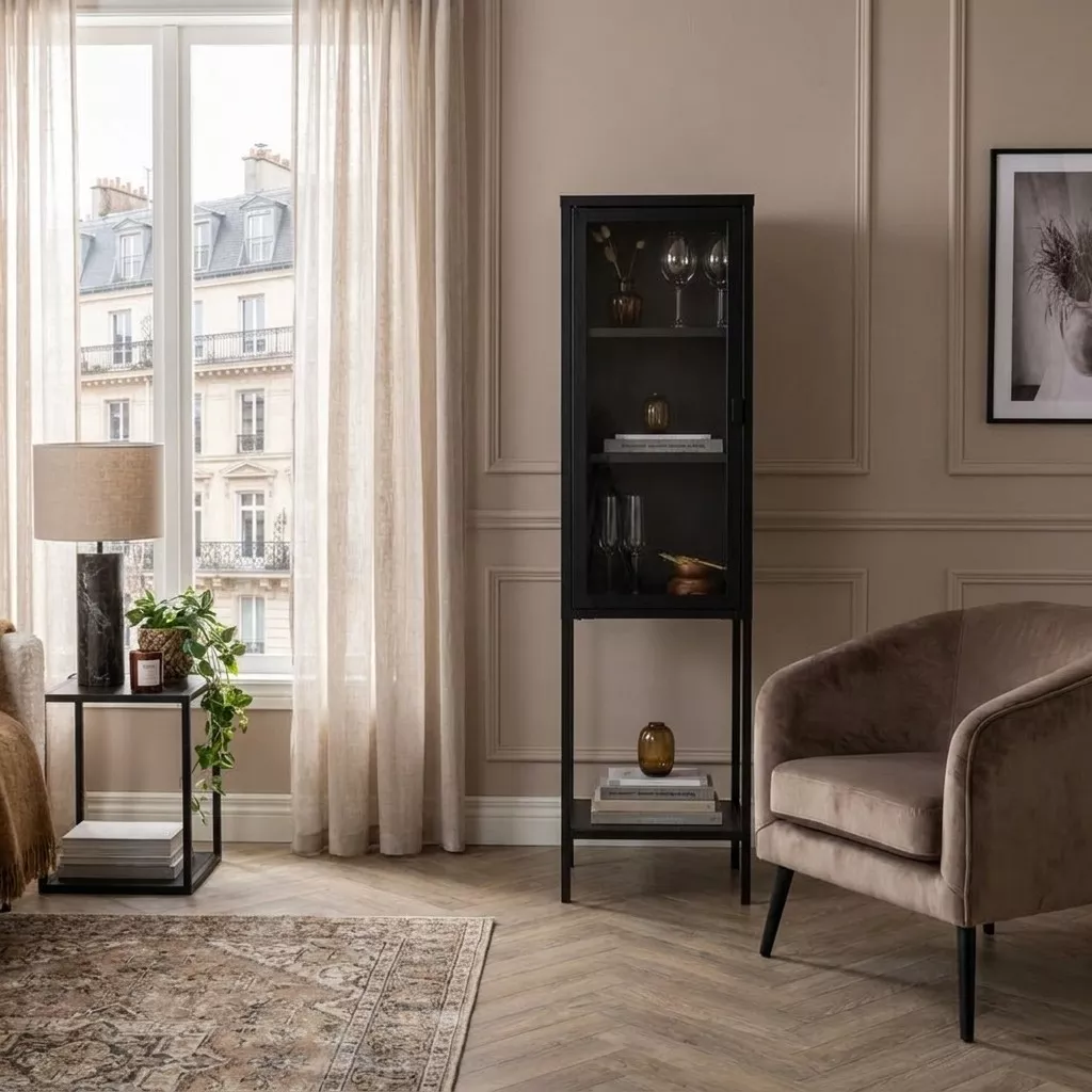 Vitrine colonne noire en acier thermolaqué et verre clair, posée contre une boiserie beige dans un séjour parisien.