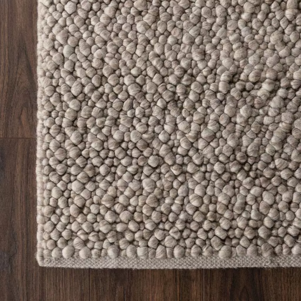 Détail macro du coin du tapis bouclette beige grège, boucles denses laine et viscose, liseré plat ton sur ton, parquet noyer