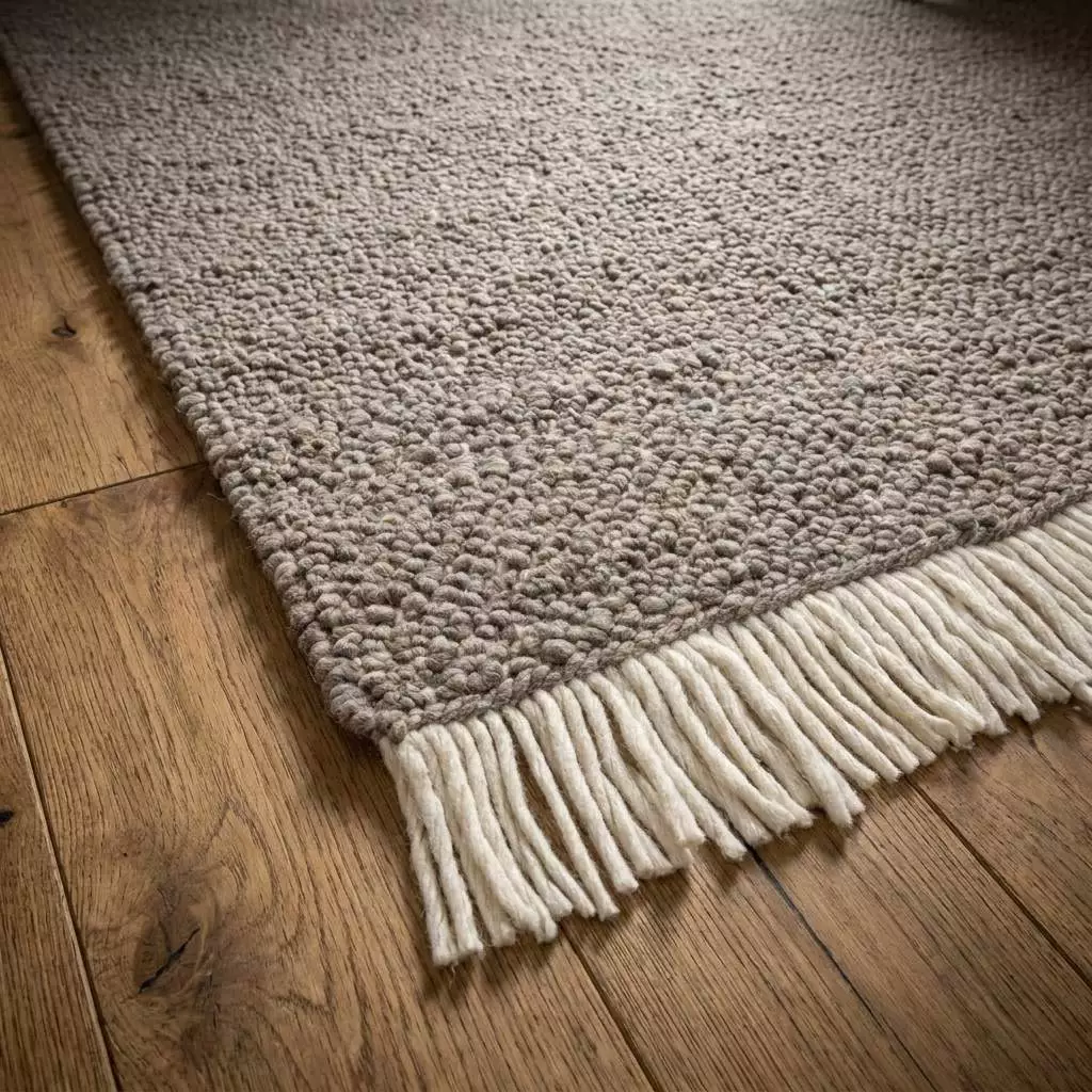 Coin du tapis laine bouclé greige vu en angle rasant, boucles serrées et franges écrues épaisses sur parquet chêne clair