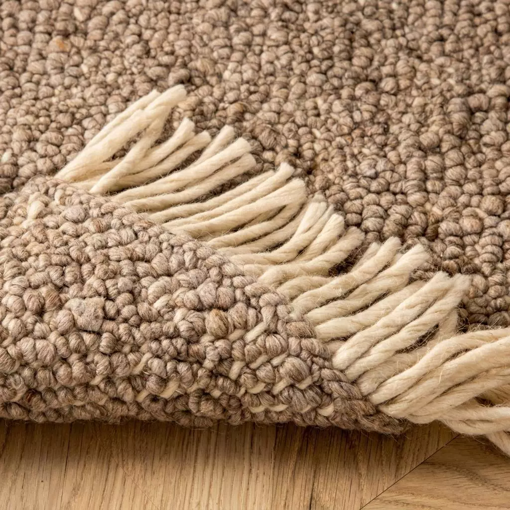 Gros plan sur les boucles serrées du tapis laine greige et les franges écrues torsadées, coin retourné révélant la trame