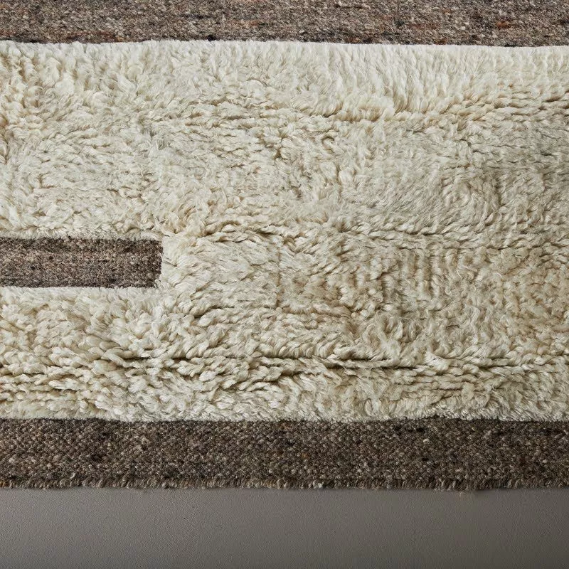 Gros plan zone shaggy laine ivoire bouclée, relief dense et moelleux, transition irrégulière vers bande kilim gris chiné