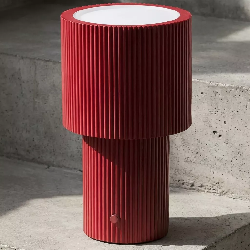 Lampe à poser rechargeable rouge cannelée vue de près sur un sol en béton brut, lumière naturelle latérale et fond minéral.