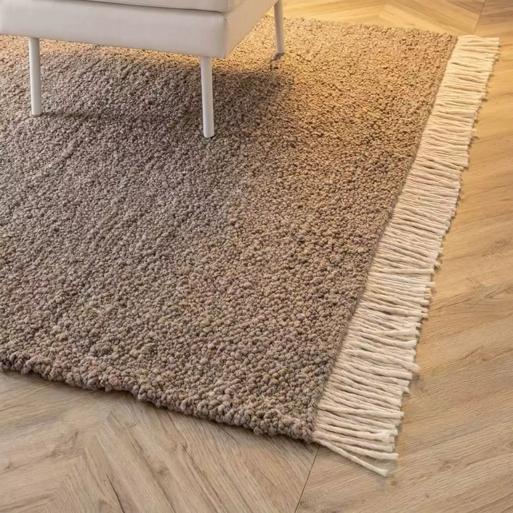 Coin du tapis laine bouclé greige en contexte, texture dense et franges écrues visibles, parquet chêne à chevrons en arrière-plan