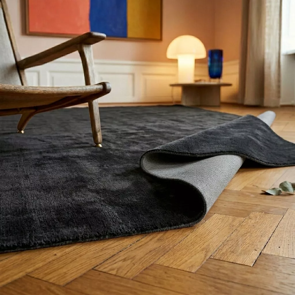 Tapis viscose anthracite avec coin replié montrant l'envers en toile, fauteuil bois clair et lampe champignon opaline