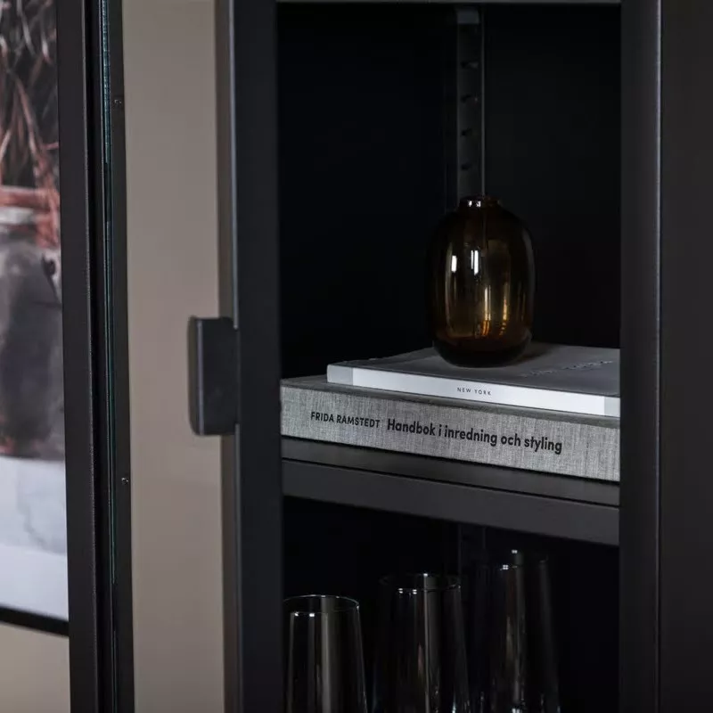 Détail intérieur de la vitrine colonne noire en acier thermolaqué : étagère avec verres à pied, vase et livre.