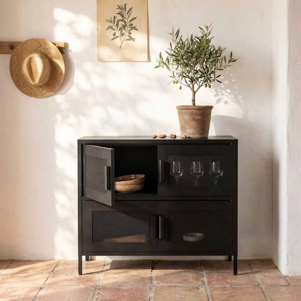 Buffet bas en acier noir mat à portes en grillage losange contre un mur blanc, petit arbre en pot et sol en terre cuite