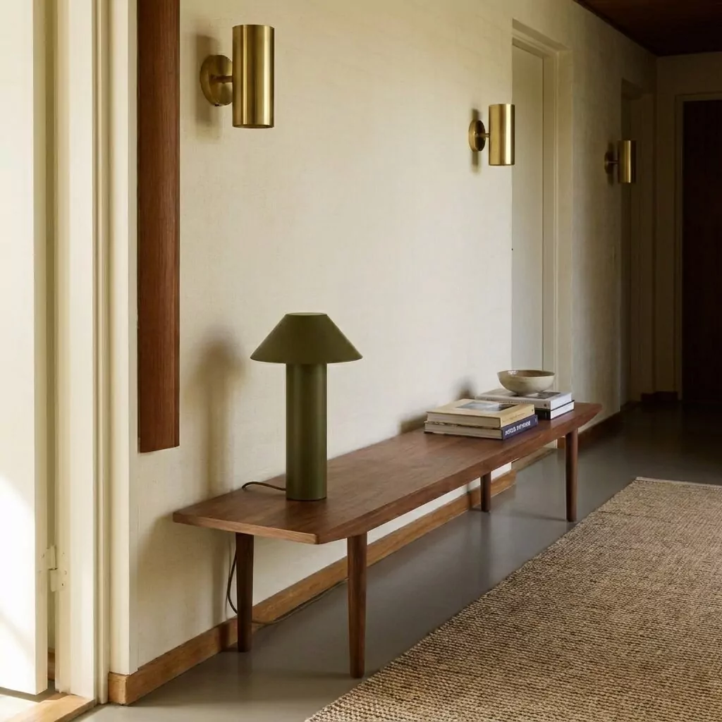 Lampe à poser vert olive mat sur un banc en bois foncé, appliques tubulaires dorées au mur, esprit mid-century