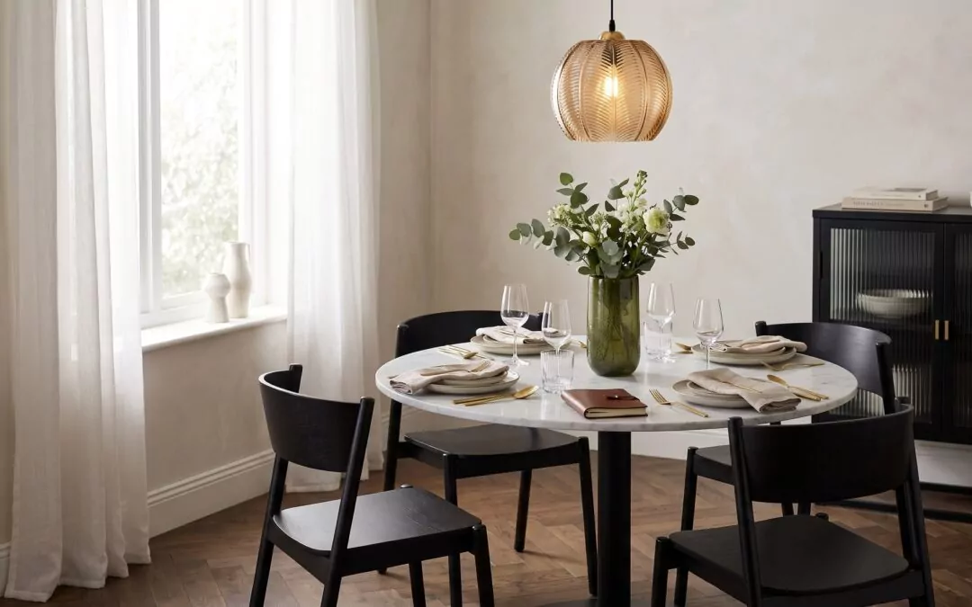 Salle à manger graphique : table ronde en marbre blanc et pied noir, chaises noires galbées, suspension en verre ambré et laiton, buffet noir cannelé, vase vert olive, parquet chêne.