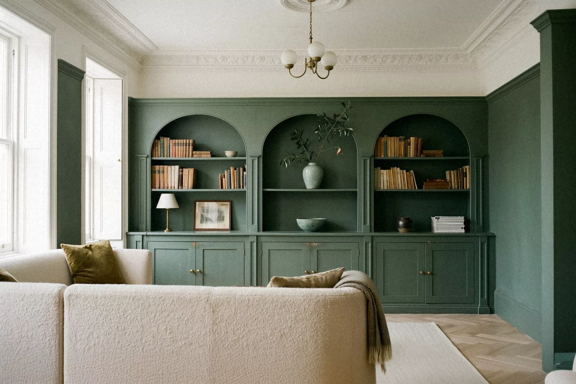 Salon bibliothèque vert sauge dans une maison de ville, boiseries à arches peintes du sol au plafond, canapé en laine bouclée écrue, lustre à globes opalins, corniche moulurée et parquet à chevrons